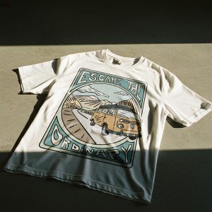 T-shirt bianca con grafica illustrata di un furgoncino giallo su una strada tra le montagne e scritta “Escape the Ordinary”, appoggiata a terra nella luce del sole