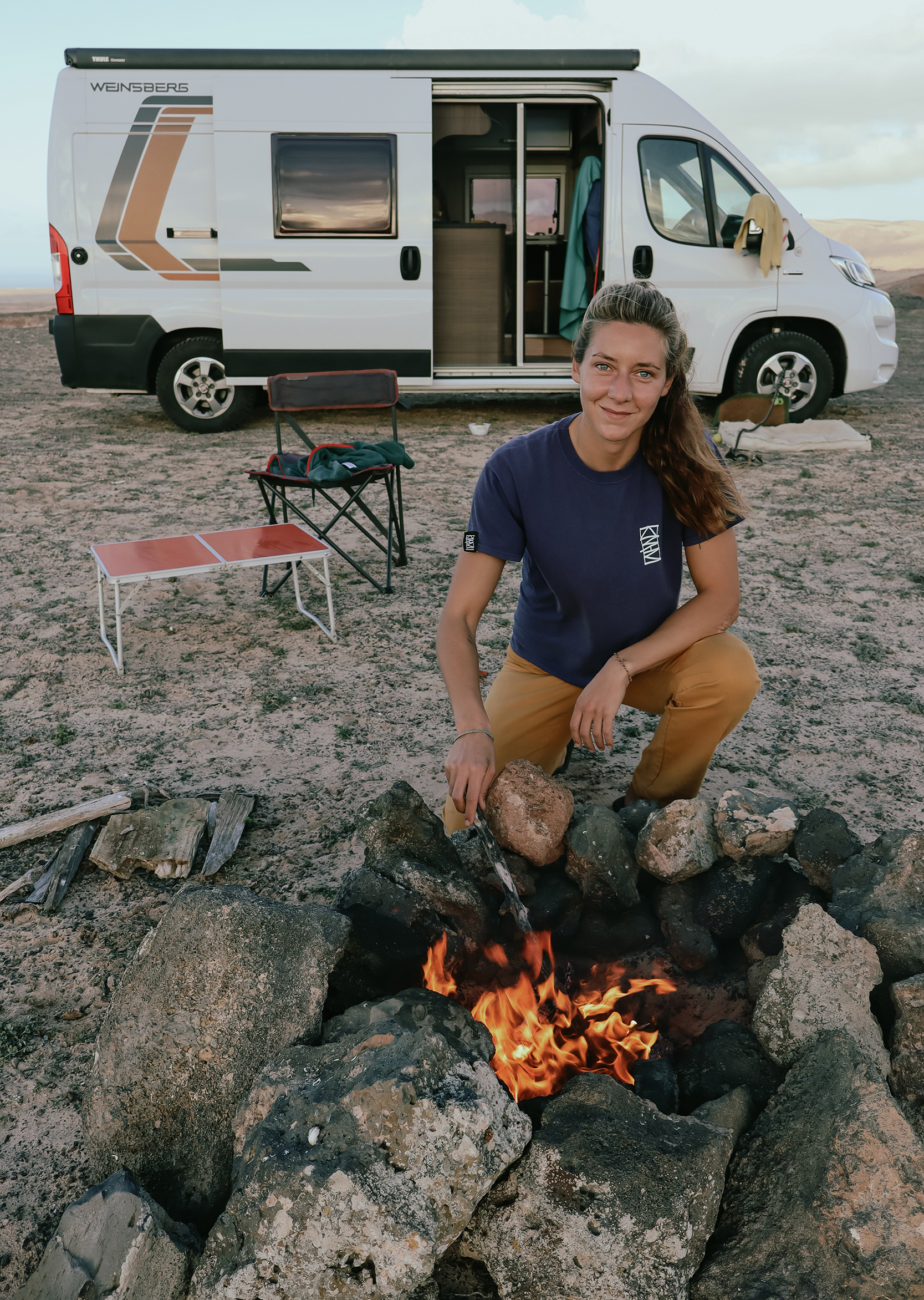 Foto di Abir Nazha che indossa una maglia di athenaabir davanti a un fuoco acceso per grigliare con dietro il suo camper weinsberg ducato fiat. Paesaggio desertico in mezzo a rocce laviche.